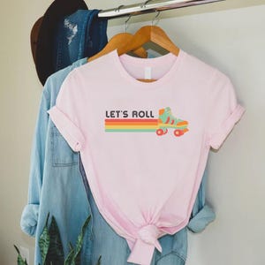 May include: Pink t-shirt featuring a retro roller skate graphic and the text "Let's Roll" in black.