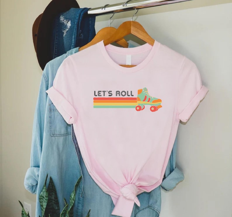 May include: Pink t-shirt featuring a retro roller skate graphic and the text "Let's Roll" in black.