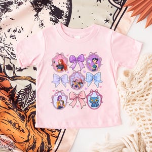 May include: A light pink toddler t-shirt featuring a design of colorful bows and framed illustrations of cartoon characters. The illustrations include a cat, a group of people, and individual portraits. The shirt is made of a soft material.