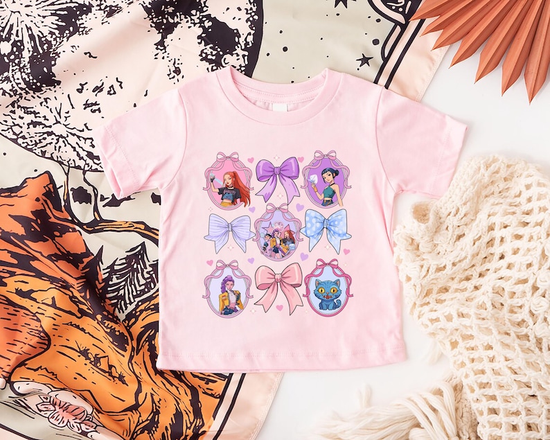 May include: A light pink toddler t-shirt featuring a design of colorful bows and framed illustrations of cartoon characters. The illustrations include a cat, a group of people, and individual portraits. The shirt is made of a soft material.