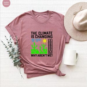 May include: A dusty rose t-shirt with the text "THE CLIMATE IS CHANGING WHY AREN'T WE?" and a graphic of a sun, thermometer, and cacti. A white mug and a beige hat are next to the shirt.