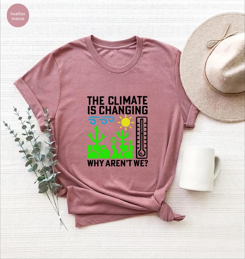 May include: A dusty rose t-shirt with the text "THE CLIMATE IS CHANGING WHY AREN'T WE?" and a graphic of a sun, thermometer, and cacti. A white mug and a beige hat are next to the shirt.