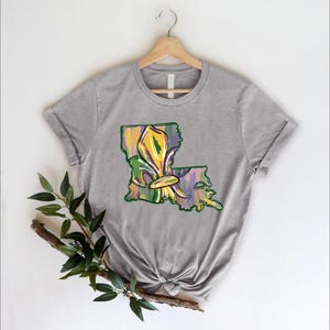 May include: A gray t-shirt featuring a colorful graphic of the state of Louisiana with a fleur-de-lis design. The graphic incorporates yellow, green, and purple hues, evoking a festive atmosphere. The shirt is displayed on a wooden hanger.