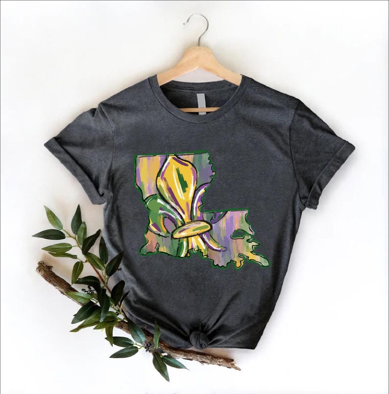 May include: Dark gray t-shirt featuring a colorful fleur-de-lis design within the outline of Louisiana. The design incorporates yellow, green, and purple hues, evoking a festive and artistic aesthetic. The shirt is displayed on a wooden hanger.