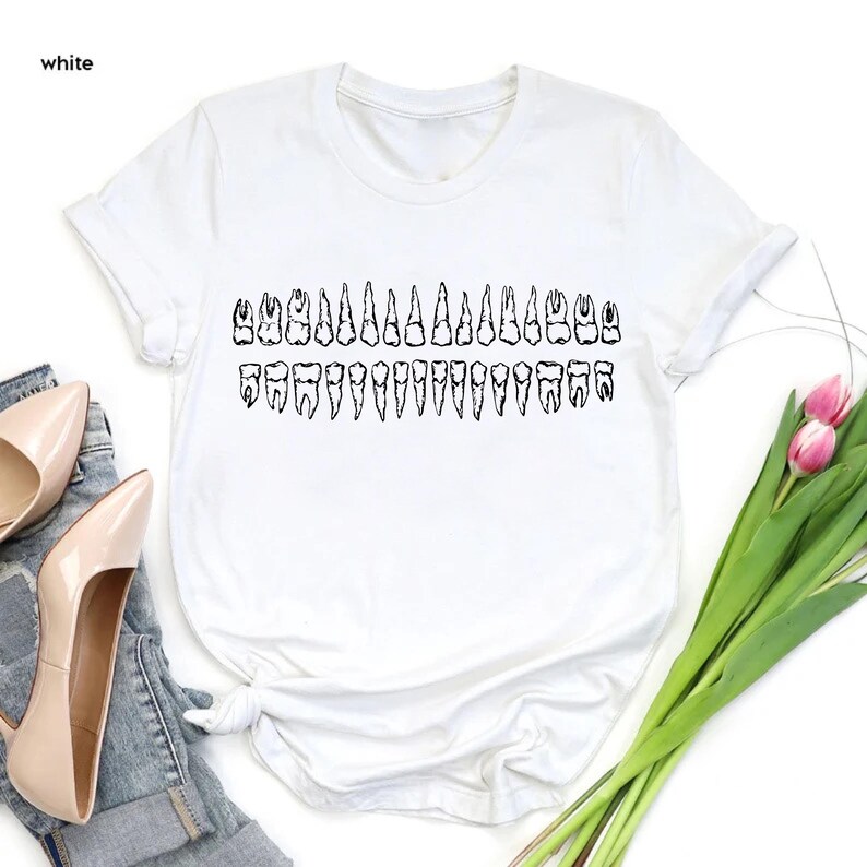 May include: White t-shirt featuring a black graphic of a full set of teeth. The text "white" is in the upper left corner. The shirt is styled with a pair of beige heels and a pair of blue jeans. A bouquet of pink tulips is in the background.