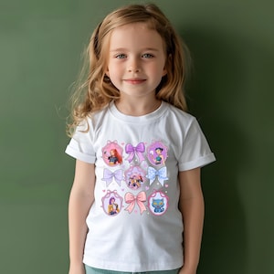 May include: White t-shirt featuring a colorful graphic design of cartoon characters in oval frames with bows. The design includes pink, purple, and blue bows, and the characters have various hair colors. The shirt is worn by a young girl.