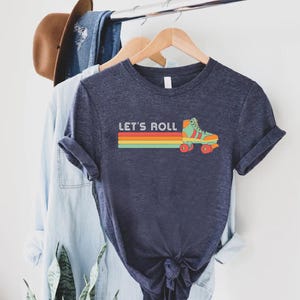 May include: A navy blue t-shirt featuring a retro rainbow stripe and a vintage roller skate graphic. The text "Let's Roll" is printed on the shirt.