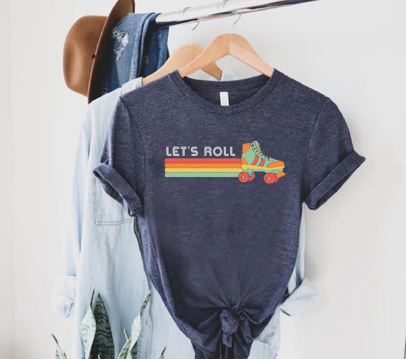 May include: A navy blue t-shirt featuring a retro rainbow stripe and a vintage roller skate graphic. The text "Let's Roll" is printed on the shirt.