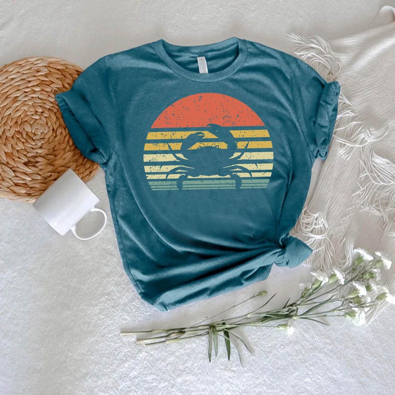 May include: Teal-colored t-shirt featuring a crab silhouette set against a retro sunset design. The sunset has horizontal stripes in orange, yellow, and green. The shirt is a casual, short-sleeved top, perfect for beach or summer wear.