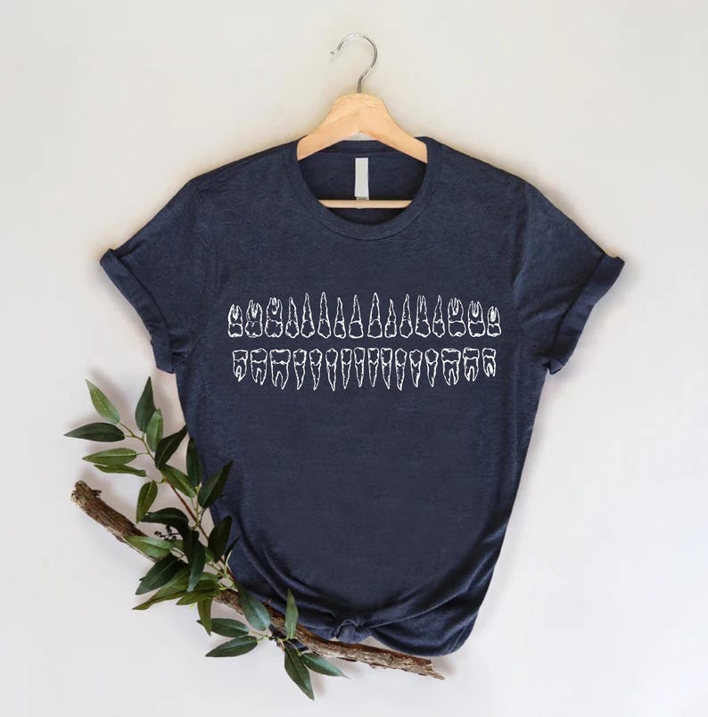 May include: Navy blue t-shirt featuring a white graphic of a complete set of human teeth. The teeth are arranged in two rows, representing the upper and lower jaws. The shirt is displayed on a wooden hanger against a white background.