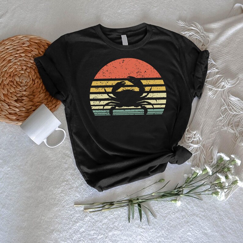 May include: Black t-shirt featuring a crab silhouette against a retro sunset design. The sunset has horizontal stripes in orange, yellow, and teal. The shirt is made of a soft material and has short sleeves.