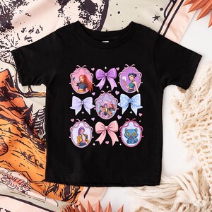 May include: Black t-shirt featuring a colorful graphic design. The design includes illustrations of characters in oval frames with bows. The bows are in various colors, including pink, purple, and blue. The shirt is made of a soft material.