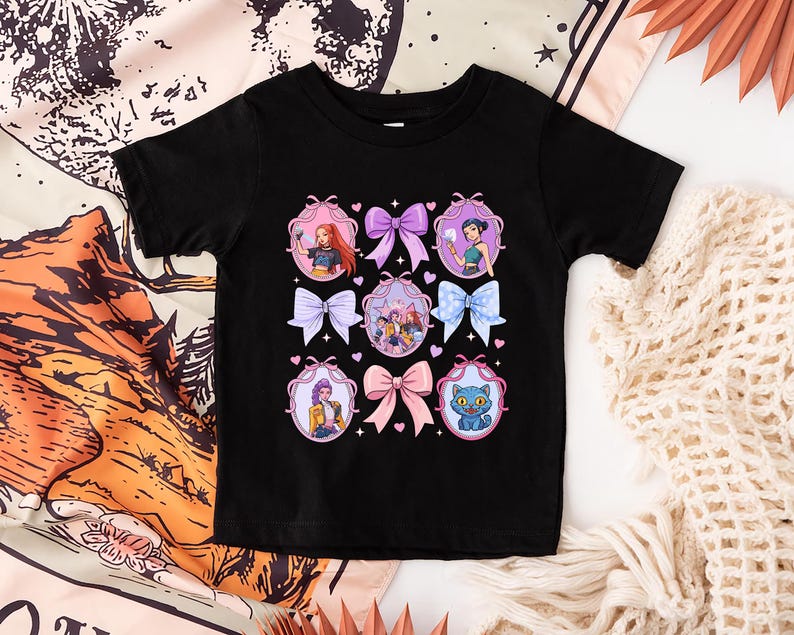 May include: Black t-shirt featuring a colorful graphic design. The design includes illustrations of characters in oval frames with bows. The bows are in various colors, including pink, purple, and blue. The shirt is made of a soft material.