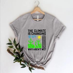 May include: A gray t-shirt with the text "THE CLIMATE IS CHANGING WHY AREN'T WE?" in black. The design includes a sun, thermometer, and green cacti. The shirt is hanging on a wooden hanger.