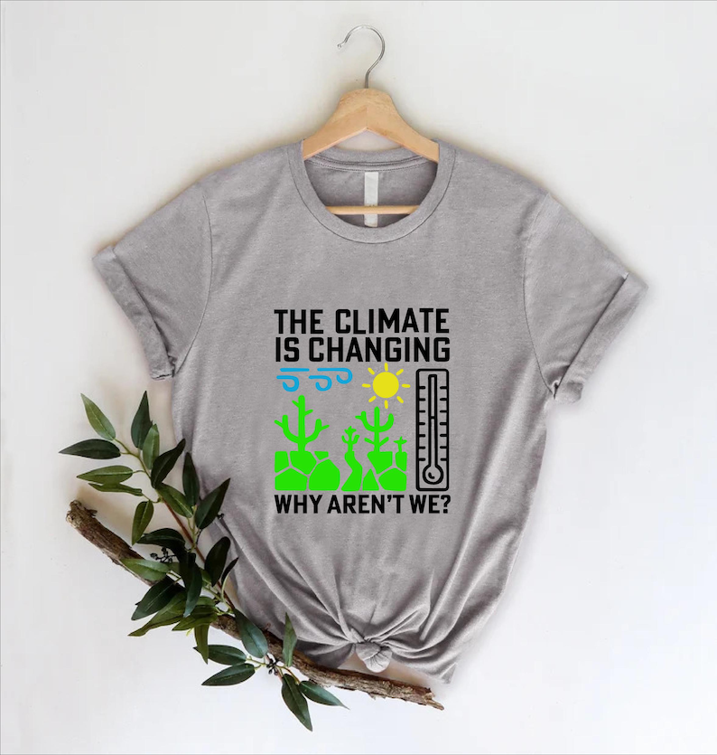 May include: A gray t-shirt with the text "THE CLIMATE IS CHANGING WHY AREN'T WE?" in black. The design includes a sun, thermometer, and green cacti. The shirt is hanging on a wooden hanger.