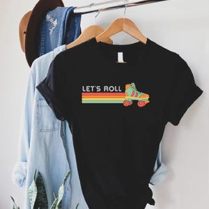 May include: Black t-shirt featuring a retro roller skate graphic and the text "Let's Roll" in white. The roller skate is green, orange, and yellow with red wheels. The text is in a white sans-serif font.