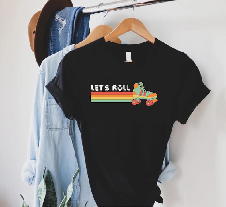May include: Black t-shirt featuring a retro roller skate graphic and the text "Let's Roll" in white. The roller skate is green, orange, and yellow with red wheels. The text is in a white sans-serif font.