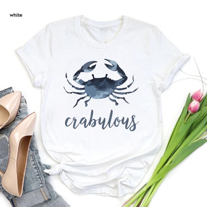 以下が含まれることがあります： 青いカニのイラストと「crabulous」の文字が入った白いTシャツ。カニのイラストは水彩画風です。ジーンズとハイヒールでスタイリングされています。