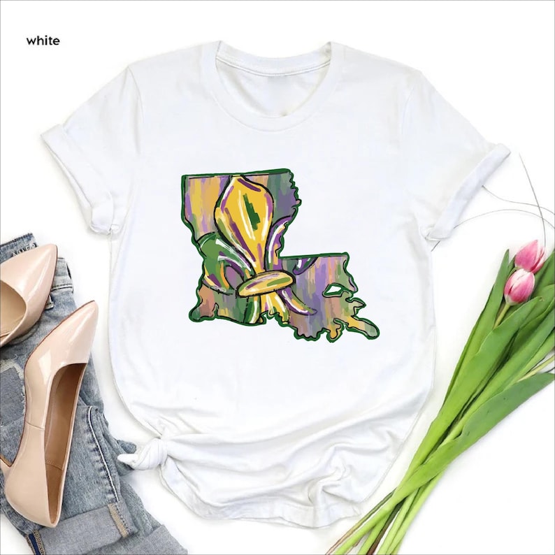 May include: White t-shirt featuring a colorful design of the Louisiana state outline with a fleur-de-lis. The graphic incorporates shades of green, yellow, and purple, evoking a festive and artistic vibe. The shirt is a casual wear item.