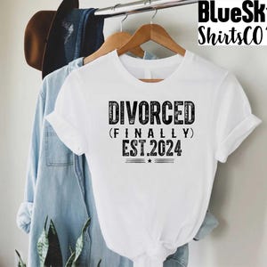May include: White t-shirt with the text "DIVORCED (FINALLY) EST.2024" in a distressed black font. The shirt is hanging on a wooden hanger. A denim shirt and a brown hat are visible in the background.