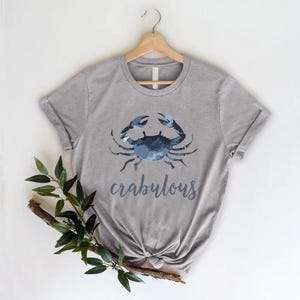 以下が含まれることがあります： 青いカニのグラフィックと「crabulous」の文字がスクリプトフォントでプリントされたグレーのTシャツ。木製のハンガーに掛けられています。白い背景に、左下隅に緑の葉と枝があります。