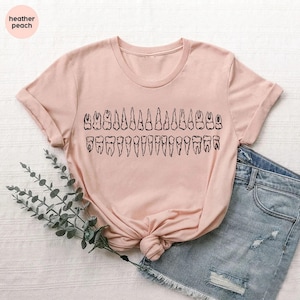 May include: A light pink t-shirt featuring a black graphic of a set of teeth. The shirt has short sleeves and a crew neck. The text "heather peach" is in a circle in the upper left corner.