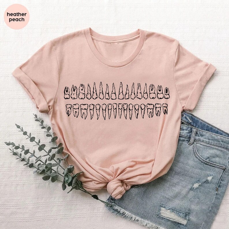 May include: A light pink t-shirt featuring a black graphic of a set of teeth. The shirt has short sleeves and a crew neck. The text "heather peach" is in a circle in the upper left corner.