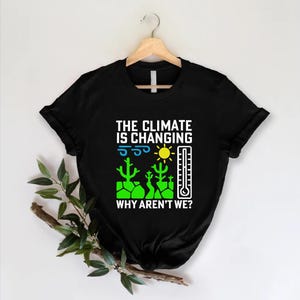 May include: Black t-shirt with the text "THE CLIMATE IS CHANGING WHY AREN'T WE?" in white. The graphic includes a sun, thermometer, cacti, and wind symbols in green, yellow, and blue. The shirt is hanging on a wooden hanger.