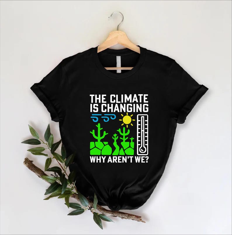 May include: Black t-shirt with the text "THE CLIMATE IS CHANGING WHY AREN'T WE?" in white. The graphic includes a sun, thermometer, cacti, and wind symbols in green, yellow, and blue. The shirt is hanging on a wooden hanger.