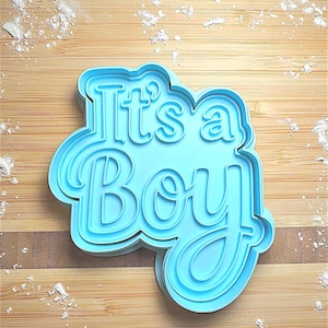 May include: A light blue cookie cutter with the phrase "It's a Boy" in a layered, cursive font. The cutter is on a wooden surface with scattered white powder, likely flour or powdered sugar.