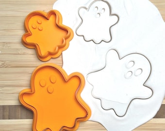 May include: Orange plastic ghost-shaped cookie cutters next to a white dough cutout. The cutters have three circular eye indentations. The dough has two ghost shapes pressed into it. The background is a light brown wooden surface.