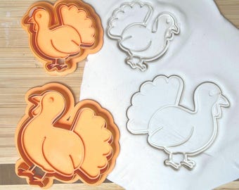 May include: Orange plastic turkey-shaped cookie cutters next to a white surface with turkey imprints. The cutters are in the shape of a turkey with a fanned tail. The turkey cookie cutters are on a wooden surface.