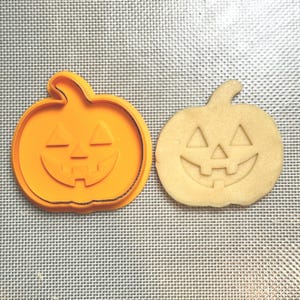 May include: An orange pumpkin-shaped cookie cutter next to a baked cookie with the same design. The cutter is made of plastic and has a smiling jack-o'-lantern face. The cookie is light brown and has the same carved face.