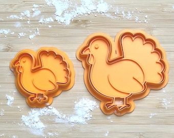 May include: Two orange plastic turkey-shaped cookie cutters. One is smaller than the other. The cutters are on a light brown wooden surface with white powder scattered around them. The turkey design includes a head, body, legs, and a fanned tail.