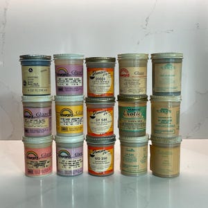 May include: A collection of 15 small glass jars of glaze for pottery. The jars are labeled with different colors and names, such as "Sandstar", "Raven Black", "Cocoa Marble", "Brocade Satin", "Oriental Green", and "Mahogany".