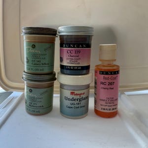 May include: Five bottles of paint, two are brown, one is gray, one is red, and one is orange. The bottles are labeled with the names of the colors and the brand names Duncan and Mayco. The bottles are all 2 fluid ounces (59 ml).