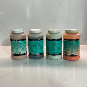 May include: Four jars of Duncan Envision Glaze in various colors, including beige, gray, light green, and orange. Each jar has a white lid and a teal label with product information. The jars are arranged on a white surface.
