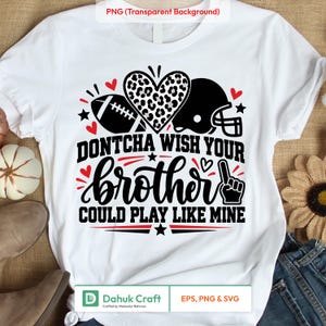Pode incluir: Uma t-shirt branca com um design gráfico preto que inclui um coração com padrão de leopardo, uma bola de futebol americano, um capacete e um dedo de espuma. O texto diz "DONTCHA WISH YOUR brother COULD PLAY LIKE MINE" em letras pretas e vermelhas.