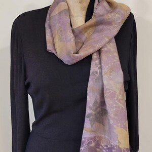 May include: A sheer scarf with a floral pattern in shades of lavender, beige, and gray. The scarf is draped over a black garment on a mannequin. The scarf's design features botanical elements.