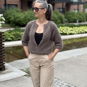 Puede incluir: Una rebeca de punto marrón con un patrón de rombos texturizado, mangas tres cuartos y cuello redondo con pequeños botones metálicos en la parte delantera, sobre una camiseta negra sin mangas con pantalones beige y gafas de sol oscuras.