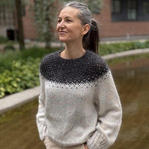Könnte beinhalten: Ein langärmeliger Strickpullover mit Rundhalsausschnitt und zweifarbigem Design. Die obere Passe ist dunkel anthrazitfarben mit einem dezenten Strukturmuster und geht in einen hellen, cremefarbenen Körper mit langen Ärmeln und gerippten Bündchen über.