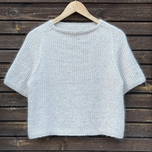 May include: A light gray, short-sleeved sweater hangs on a wooden hanger. The sweater has a crew neck and a textured knit pattern. The background is a wooden wall with horizontal planks.