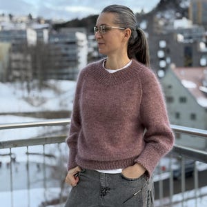 以下が含まれることがあります： モーヴ色のVネックセーターと長袖。グレーのデニムジーンズと合わせています。メガネをかけ、雪景色を背景にしたバルコニーに立っています。