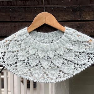 May include: A light blue knitted collar with a scalloped edge and openwork design, hanging on a wooden hanger. The collar is made of soft yarn and features a delicate, lacy pattern. The background includes a wooden wall and a white railing.