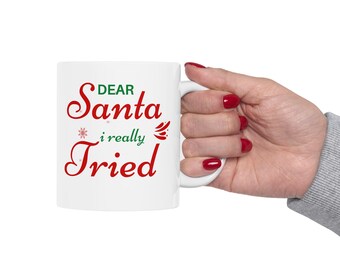 May include: White ceramic mug with the text "DEAR Santa i really Tried" in red and green. The mug has a white handle and is held by a hand with red nail polish. The mug is a Christmas-themed gift.