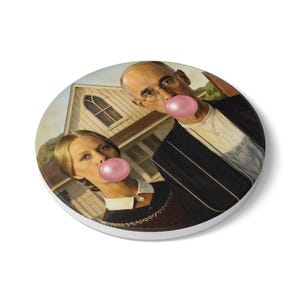 Pode incluir: Base redonda com uma paródia da pintura "American Gothic". A imagem mostra um homem e uma mulher com bolhas de chiclete rosa, em um fundo de uma casa. A base tem uma borda branca. Diâmetro de aproximadamente 10 cm.