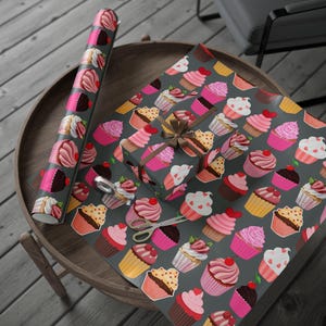 Puede incluir: Un rollo de papel de regalo con estampado de cupcakes y una caja de regalo envuelta sobre una mesa de madera. El papel y la caja presentan un fondo gris oscuro con ilustraciones coloridas de cupcakes. Un par de tijeras descansa sobre el papel.
