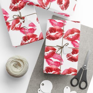 May include: Gift-wrapped presents with a white background and a pattern of red lipstick kiss marks. The gifts are tied with twine and adorned with white heart-shaped tags. A ball of twine, scissors, and blank tags are also visible.