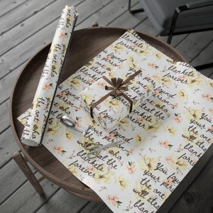May include: Gift wrapping supplies on a wooden table. The scene includes a wrapped gift with a brown ribbon, wrapping paper, and a roll of paper. The paper has a floral pattern with text that reads "You found your person...will steal the blankets forever."