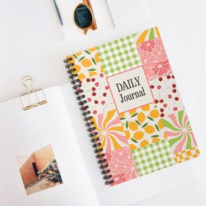 May include: A spiral-bound daily journal with a colorful patchwork design featuring lemons, cherries, and floral patterns. The cover includes the text "DAILY Journal" in a rectangular frame. The journal is open, revealing blank pages and a gold binder clip.